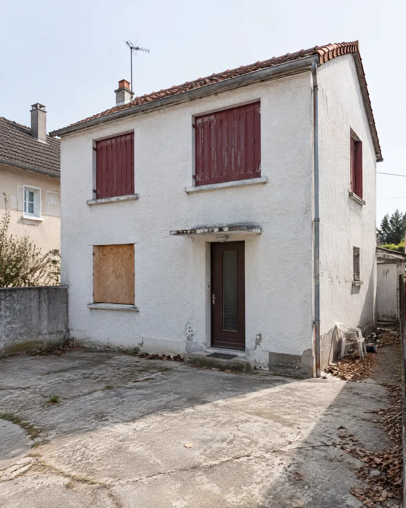 Pavillon blanc dégradé aux volets bordeaux, vu depuis une cour latérale, sans personne.